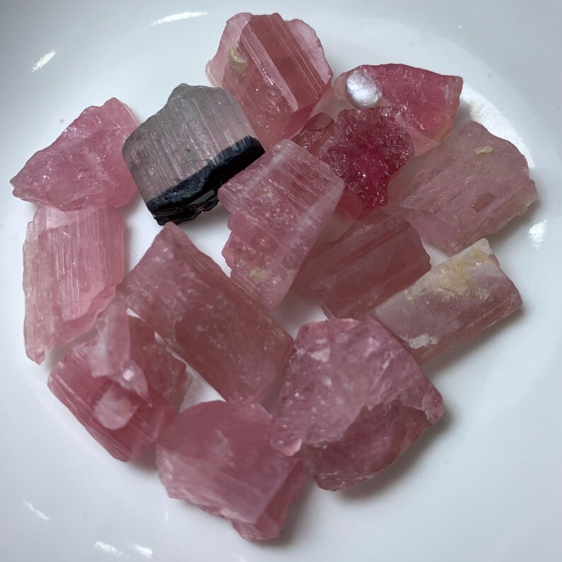 May include: A collection of pink and black tourmaline crystals. The crystals are raw and have a natural, rough texture. They are a variety of pink, from light to dark, and some have black streaks.