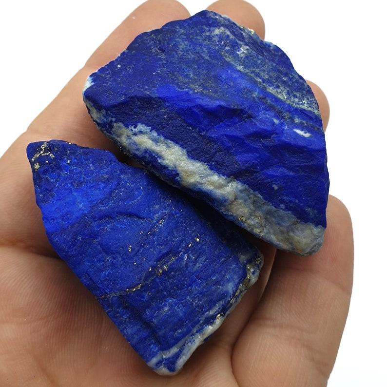 May include: Two rough, natural pieces of lapis lazuli, a deep blue gemstone with flecks of gold pyrite. The stones are irregular in shape and have a textured surface.