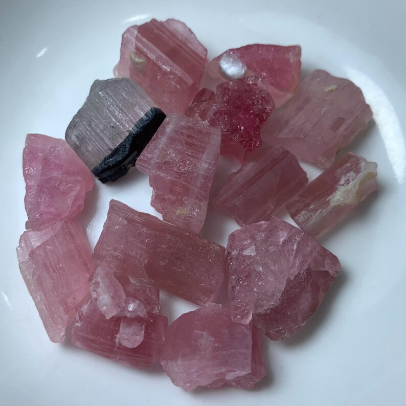 May include: A collection of pink and black tourmaline crystals. The crystals are rough and have a natural, unpolished look. They are a variety of pink, with some having a darker, more reddish hue. Some of the crystals have a black tourmaline inclusion.