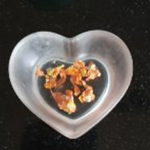 May include: A small, clear, heart-shaped dish filled with irregularly shaped, metallic gold flakes. The dish is made of a translucent material, and the flakes have a textured, uneven surface. The dish sits on a dark, reflective surface.