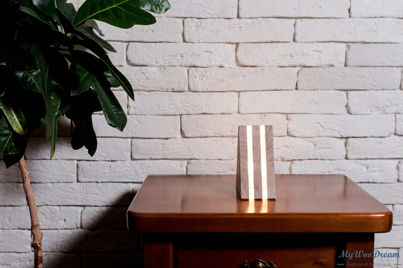 May include: A wooden lamp with a white finish and two thin, white LED lights. The lamp is sitting on a wooden table in front of a white brick wall.