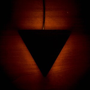 May include: A black triangular pendant light fixture hangs from a wire against a dark red wood background. The light casts a warm glow on the wood.