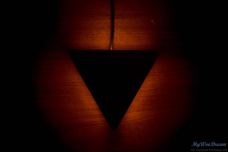 May include: A black triangular pendant light fixture hangs from a wire against a dark red wood background. The light casts a warm glow on the wood.