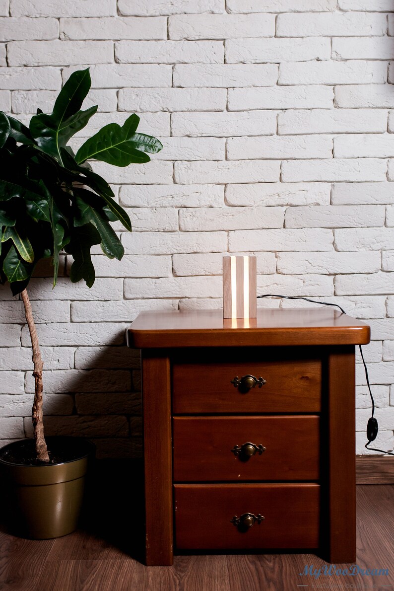 May include: A wooden lamp with a white light source sits on a wooden nightstand with three drawers. The nightstand is in front of a white brick wall. A green plant is in a brown pot to the left of the nightstand.