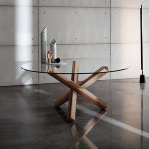 Puede incluir: Una mesa redonda de vidrio con una tapa transparente y una base de madera. La base está hecha de cuatro gruesos vigas de madera que se cruzan en el centro de la mesa. La mesa está sentada en un suelo de hormigón.