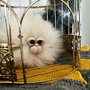 Può includere: Una scultura soffice, color crema, simile a un gatto, con grandi occhi espressivi e un naso rosa, racchiusa in una gabbia decorativa color oro. La gabbia poggia su un tessuto a righe gialle e grigie.