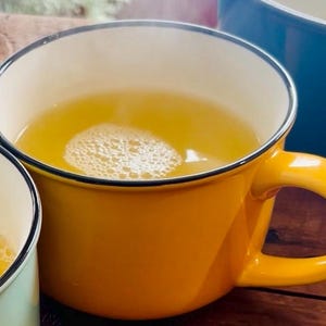 May include: A close-up of a yellow mug filled with a light yellow liquid, possibly tea or broth. The mug has a black rim and handle. Bubbles are visible on the surface of the liquid. Another mug is partially visible in the background.