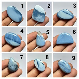 May include: Nine blue opal cabochons, each with a unique pattern and shape, are displayed on a hand. The cabochons are numbered 1 through 9.