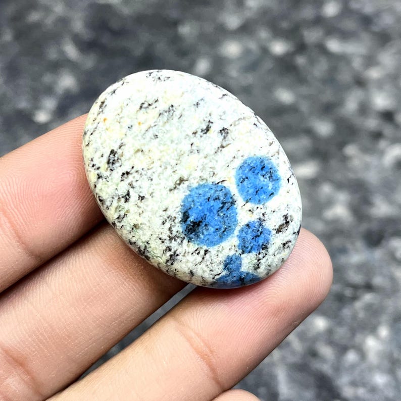 May include: Oval-shaped K2 stone, featuring a white base speckled with black and gray spots. The stone has three vibrant blue circular patterns. The polished surface of the stone is smooth and cool to the touch.