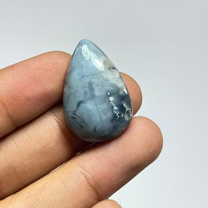 May include: A teardrop-shaped cabochon of blue opal with white and gray markings. The opal is translucent and has a smooth, polished surface.