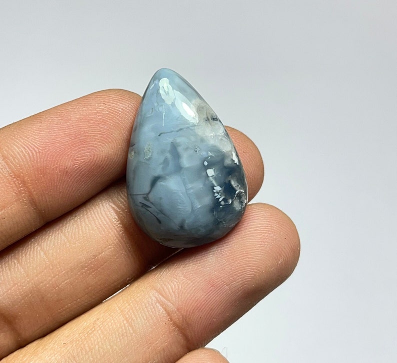 May include: A teardrop-shaped cabochon of blue opal with white and gray markings. The opal is translucent and has a smooth, polished surface.
