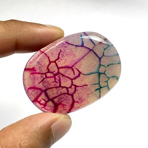 May include: An oval-shaped polished gemstone featuring a smooth, translucent surface with a distinct crackle pattern. The stone displays vibrant magenta and teal veins that contrast against a pale, cream-coloured background, creating a web-like effect.