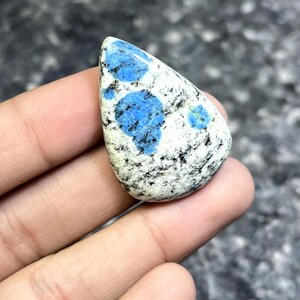 May include: A teardrop-shaped K2 stone, held in a hand, features a white base with speckled black and blue patterns. The blue patterns are concentrated in irregular shapes, creating a unique, natural design. The stone is polished and smooth.