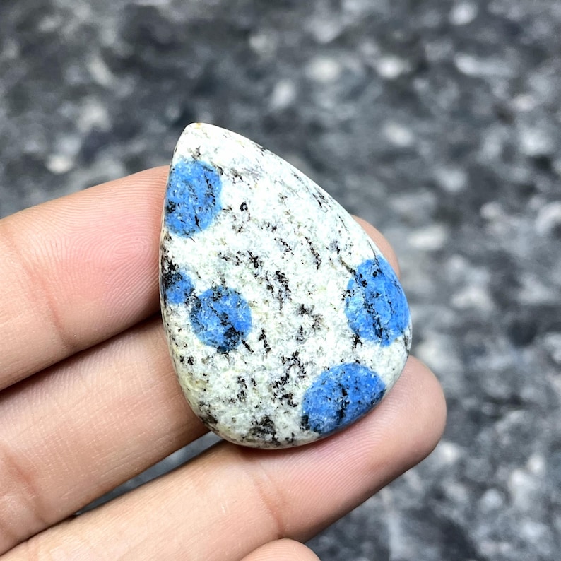 May include: A polished K2 stone, featuring a teardrop shape. The stone has a white base with black speckles and vibrant blue orb-like patterns. The gemstone is held in a hand, showcasing its unique color and texture.