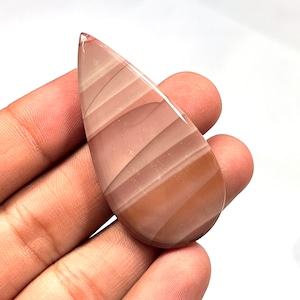 May include: A teardrop-shaped cabochon of banded agate with pink and brown stripes. The stone is translucent and has a smooth, polished surface.