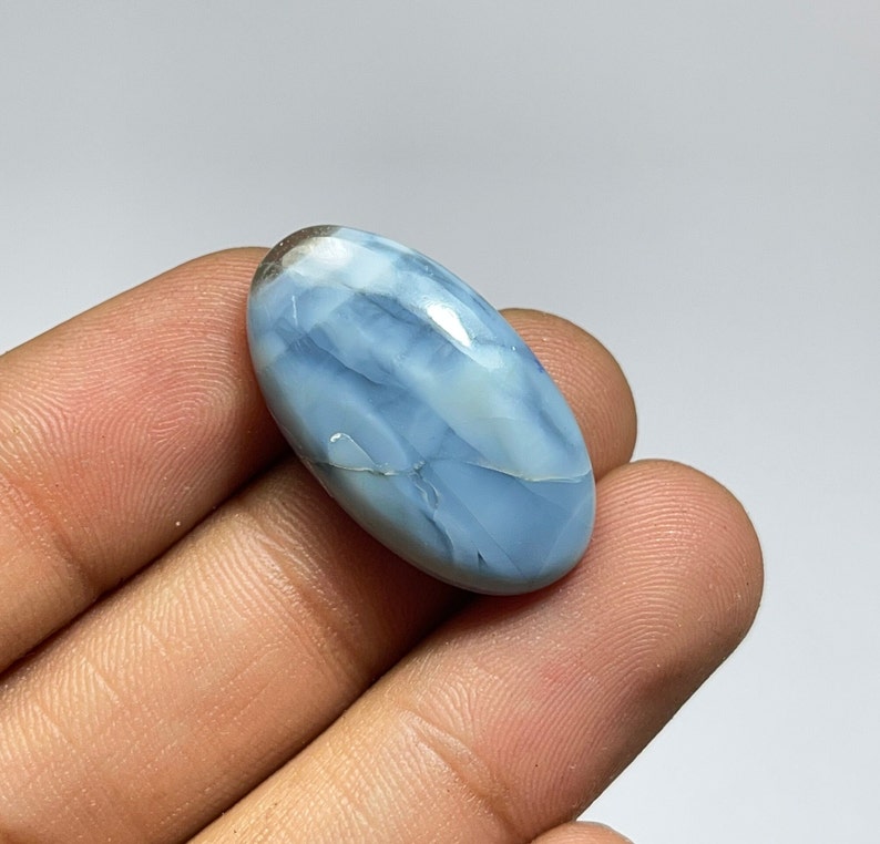 May include: A light blue oval cabochon opal gemstone with white and gray markings. The opal is smooth and polished.
