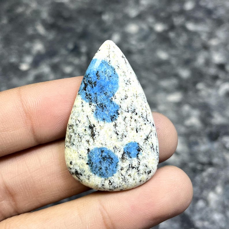 May include: A teardrop-shaped K2 stone cabochon. The stone features a creamy white base with speckled black inclusions and vibrant blue orb-like patterns. The polished surface highlights the natural textures and colors of the gemstone.