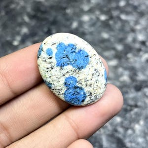 May include: Oval-shaped K2 stone, featuring a speckled white and gray base with vibrant blue patches. The polished stone is held in a hand, showcasing its natural patterns and colors. A unique gemstone for jewelry or collection.