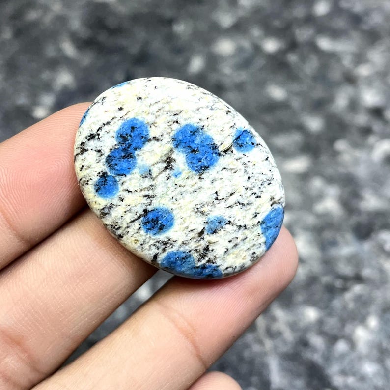 May include: Oval-shaped K2 stone with a white base, speckled with black and vibrant blue spots. The polished stone is held in a hand, showcasing its natural patterns and colors. The stone's smooth surface and unique markings make it a striking piece.