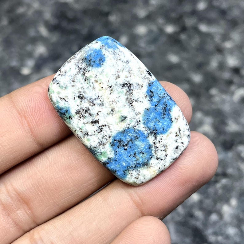 May include: A rectangular K2 stone with a polished surface. The stone features a white base with speckled black and green inclusions, and vibrant blue orb-like patterns. The stone is held in a hand.