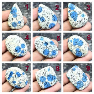 May include: Nine K2 stone cabochons, each with a unique shape and pattern. The stones feature a white base with speckled black and blue spots. The blue spots vary in size and concentration across the cabochons, offering a range of visual textures.
