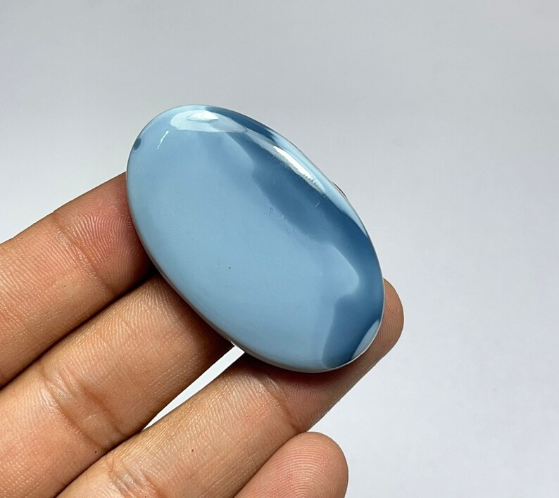 May include: A light blue oval cabochon gemstone with white and gray swirls. The gemstone is polished and has a smooth surface.