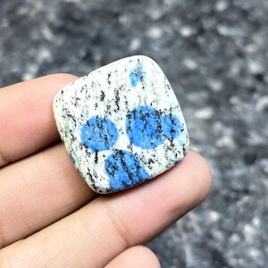 May include: A square-shaped K2 stone, featuring a white base speckled with black and blue spots. The stone has three prominent, vibrant blue circular patterns. The polished surface of the stone is smooth and reflects light.