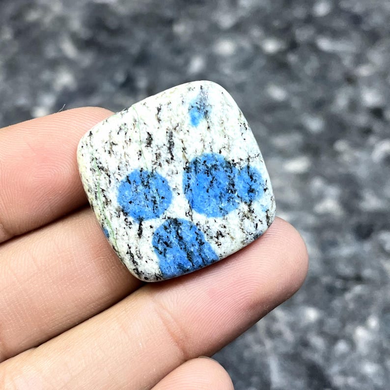 May include: A square-shaped K2 stone, featuring a white base speckled with black and blue spots. The stone has three prominent, vibrant blue circular patterns. The polished surface of the stone is smooth and reflects light.