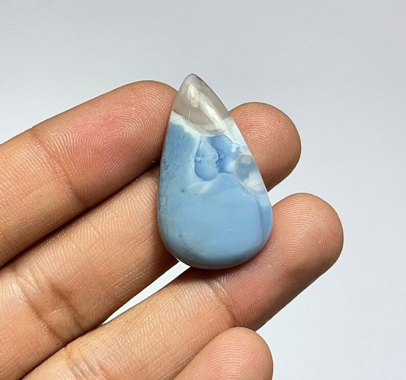 May include: A teardrop-shaped blue opal cabochon with white and gray inclusions. The opal is set in a silver tone setting.