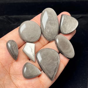 Op de afbeelding: Een verzameling van acht gepolijste zilverglans obsidiaan edelstenen in diverse vormen, waaronder ovaal, druppelvormig en driehoekig. Elke steen heeft een glad, reflecterend oppervlak met een metaalgrijze glans en subtiele interne texturen.