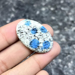 May include: An oval-shaped K2 stone, featuring a white base speckled with black and vibrant blue spots. The polished gemstone is held in a hand, showcasing its natural patterns and colors. This unique stone is a beautiful addition to any collection.