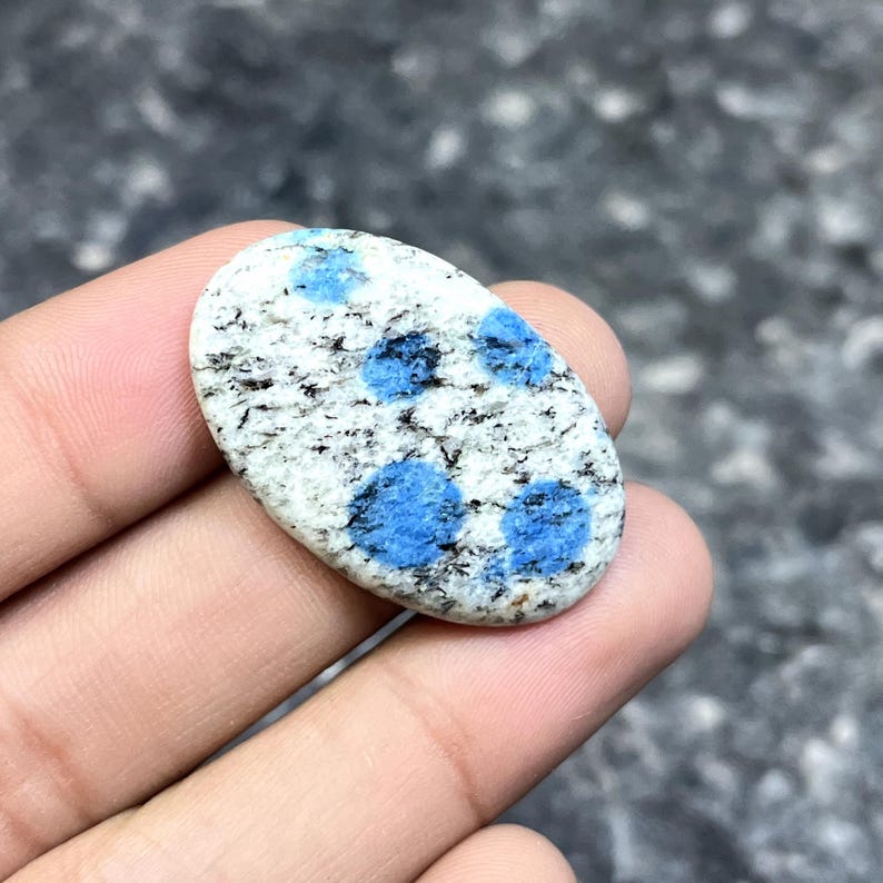 May include: An oval-shaped K2 stone, featuring a white base speckled with black and vibrant blue spots. The polished gemstone is held in a hand, showcasing its natural patterns and colors. This unique stone is a beautiful addition to any collection.
