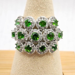 May include: A silver ring with multiple rows of round, bright green gemstones, each surrounded by smaller, clear stones. The ring is displayed on a beige, cone-shaped jewelry stand, set on a wooden base.