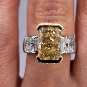 May include: A gold ring with a large, rectangular, yellow diamond in the center. The diamond is surrounded by smaller, white diamonds on either side. The ring is set in a white metal band.