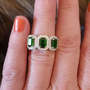 May include: A silver ring featuring three emerald-cut green gemstones, each surrounded by a halo of smaller, clear stones. The ring is displayed on a hand with orange nail polish, set against a brown, textured background.