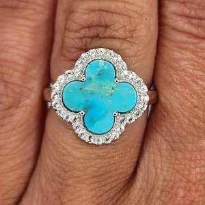 May include: A silver-toned ring featuring a four-leaf clover-shaped centerpiece with a bright turquoise stone inlay. The clover is surrounded by a halo of small, clear, round-cut stones set into the metal frame, mounted on a thin, polished band.