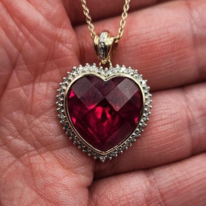 May include: A gold-toned necklace featuring a heart-shaped pendant with a faceted, deep red gemstone. The pendant is surrounded by a halo of small, clear stones. The necklace is displayed against a neutral background.
