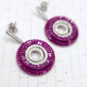 May include: A pair of silver earrings with a circular design. The earrings feature a vibrant magenta outer ring, a central spiral of white stones, and a silver post. The earrings are set against a white background.