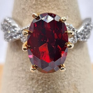 Peut inclure: Bague en or et argent avec une grosse pierre précieuse rouge foncé, facettée et de forme ovale. La bague a un sertissage en or autour de la pierre précieuse et des accents argentés sur le bandeau. La bague est exposée sur un présentoir beige.