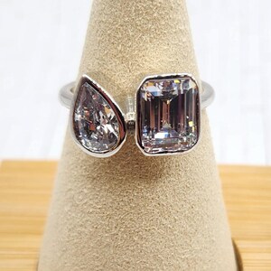May include: A silver ring with two clear gemstones. The larger gemstone is a rectangular emerald cut and the smaller gemstone is a pear shaped stone. The ring is displayed on a wooden ring holder.