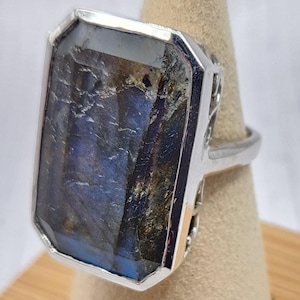 May include: A rectangular labradorite gemstone ring set in a polished silver bezel. The stone features a faceted surface with deep blue and dark grey iridescent flashes. The silver band has an intricate, openwork filigree design on the sides.