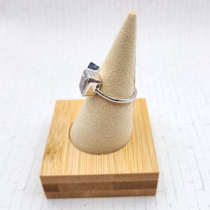 May include: A silver ring with a large, clear, square-shaped gemstone set in a bezel setting. The ring is displayed on a small, wooden ring stand.