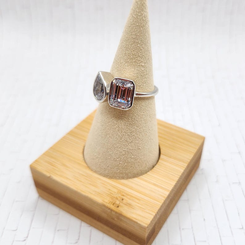 May include: A silver ring with two clear stones. The larger stone is a square emerald cut and the smaller stone is a teardrop shape. The ring is displayed on a wooden ring holder.