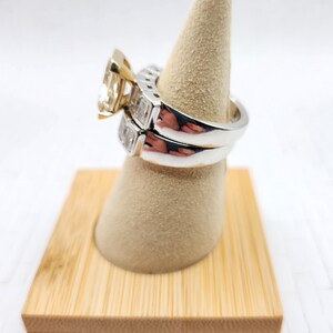 May include: A pair of engagement rings on a wooden ring display stand. The top ring features a large, square-cut diamond with a yellow gold band. The bottom ring is a simple band with smaller diamonds set in a white gold band.