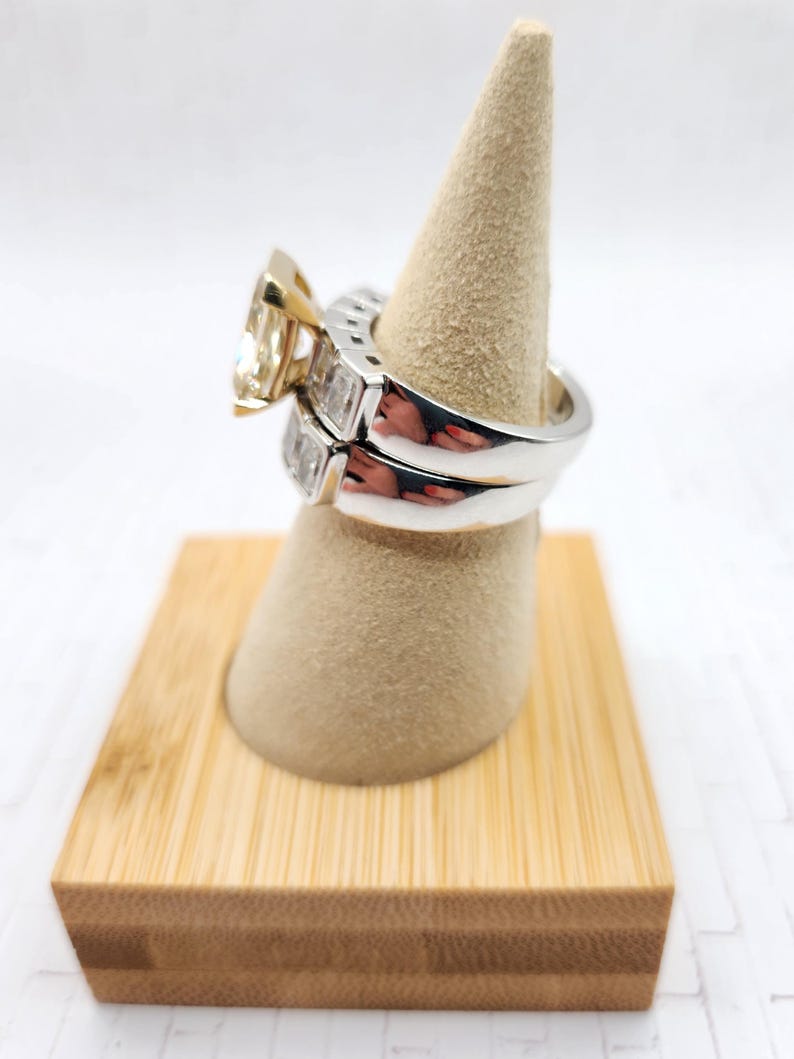 May include: A pair of engagement rings on a wooden ring display stand. The top ring features a large, square-cut diamond with a yellow gold band. The bottom ring is a simple band with smaller diamonds set in a white gold band.