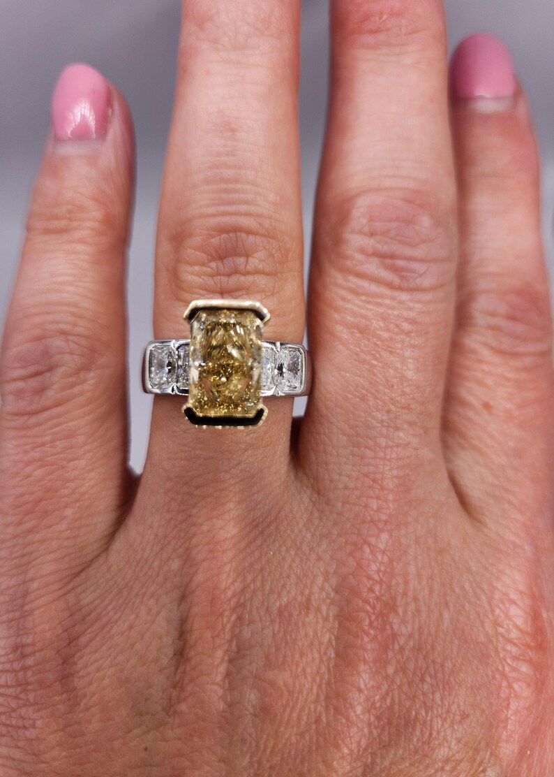 May include: A gold ring with a large, rectangular, yellow diamond in the center. The diamond is surrounded by smaller, white diamonds on either side. The ring is set in a white metal band.