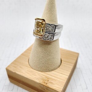 May include: A two-piece ring set with a large, square-shaped, yellow diamond center stone. The ring is set in white gold with a yellow gold band. The ring is displayed on a wooden ring holder.
