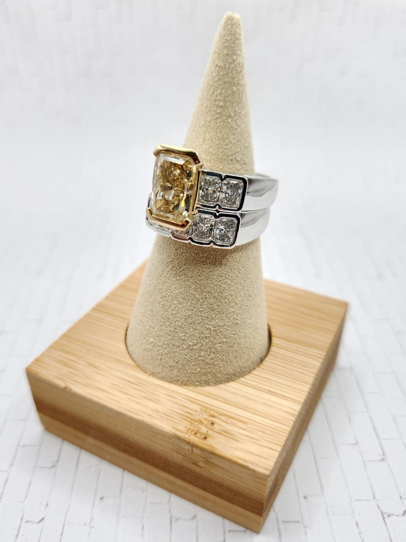 May include: A two-piece ring set with a large, square-shaped, yellow diamond center stone. The ring is set in white gold with a yellow gold band. The ring is displayed on a wooden ring holder.