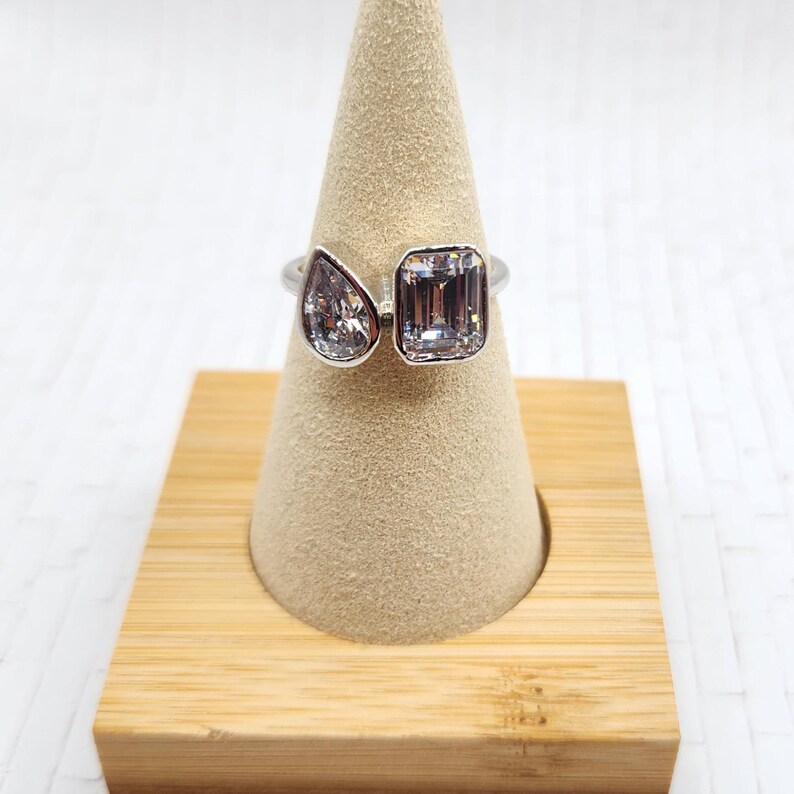 May include: A silver ring with two clear gemstones. The larger gemstone is a rectangular emerald cut and the smaller gemstone is a pear shaped stone. The ring is displayed on a wooden ring holder.