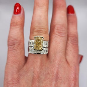 May include: A close-up of a hand wearing a ring with a large, rectangular, yellow diamond surrounded by smaller diamonds. The ring is set in white gold and has a band of smaller diamonds.